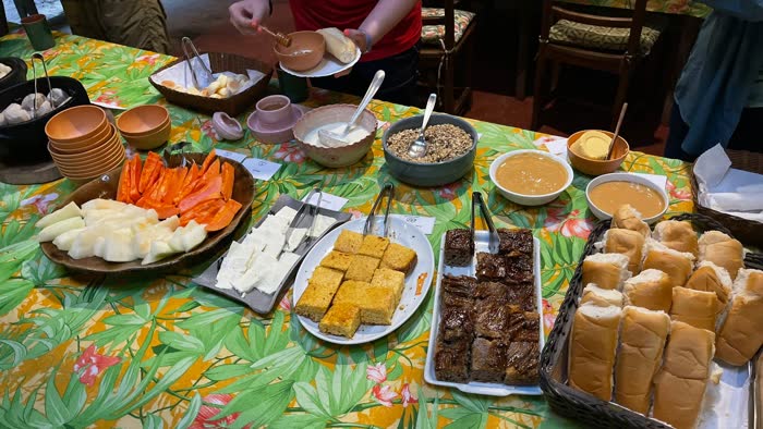 Café da manhã servido em travessas: melão e mamão fatiados, queijo fresco, bolo de fubá, bolo de banana, granola, doce de leite e cesta de pão