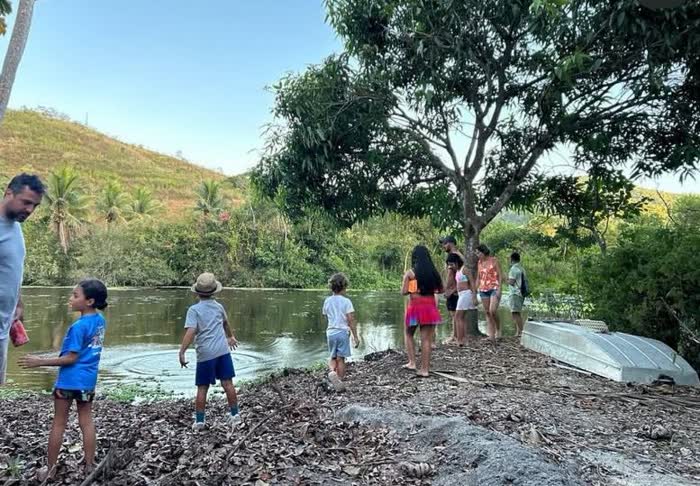 Adultos e crianças parados na margem de um lago, à sombra de uma árvore, com um barco emborcado na areia e o morro ao fundo