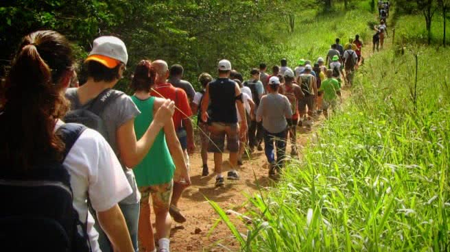 Grupo de pessoas de várias idades caminhando em fila por uma trilha de terra, entre mata e capim alto