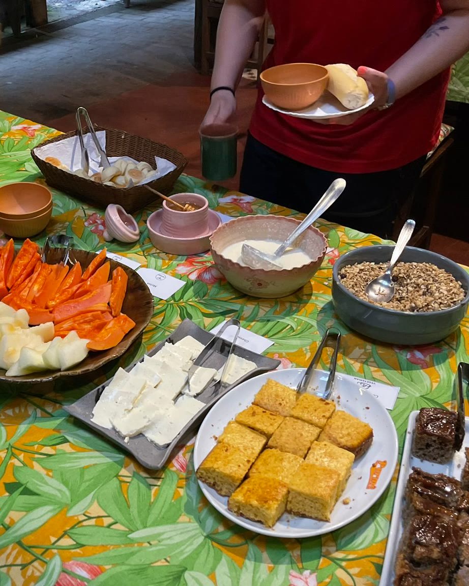 Mesa do café da manhã da fazenda com travessa de melão e mamão, queijo fresco, bolo de fubá, iogurte e granola, e alguém se servindo ao fundo